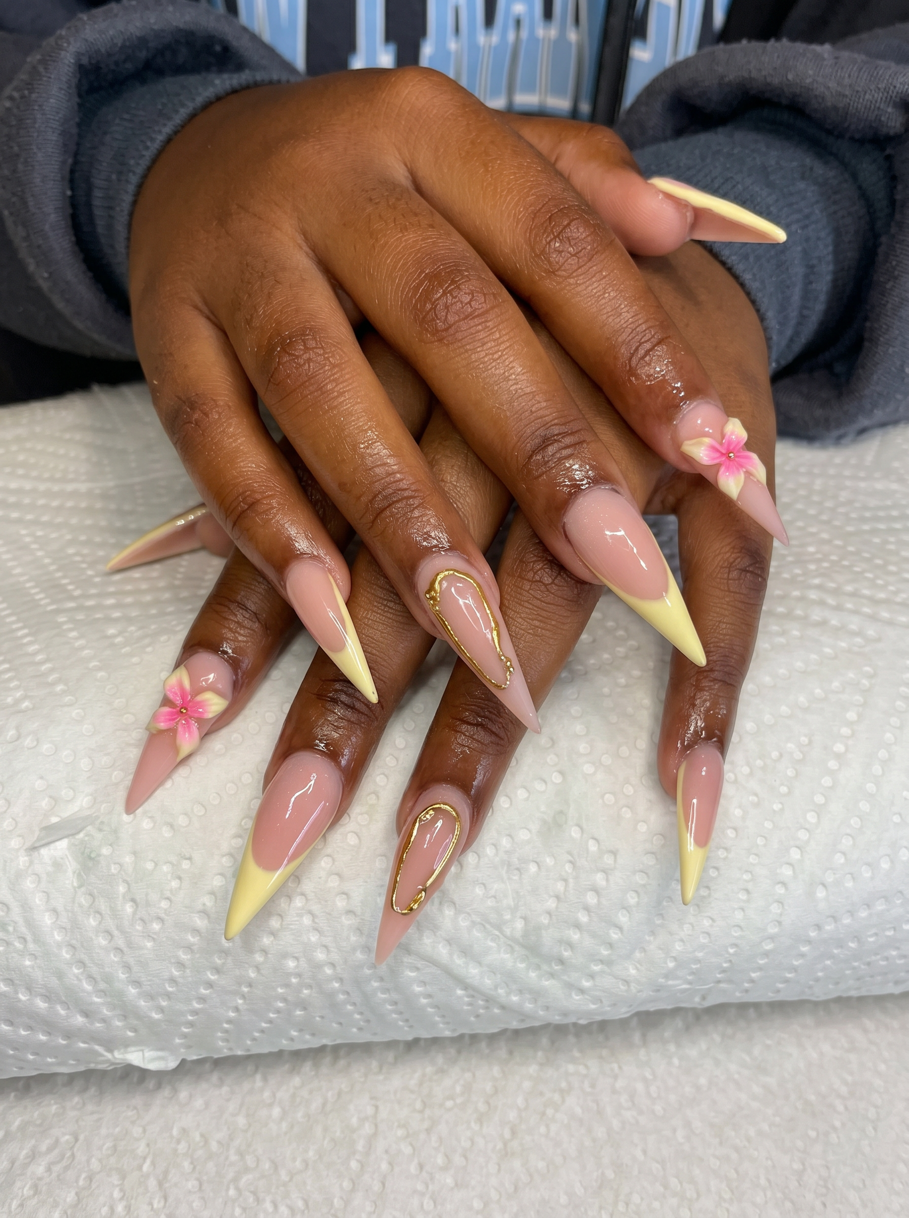Long stiletto acrylic nails with pastel yellow French tips, gold wire accents, and 3D pink flower charms by Fine Artistry
